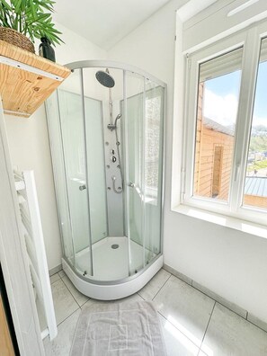 A glass-enclosed shower with a white tiled floor, a window with a view of a brick wall, and a wooden shelf with a plant. - Dance of the Waves | Sea View Balcony • Fécamp • Near Étretat (Fécamp)