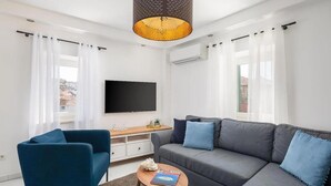 Ett modernt vardagsrum med en grå soffa, en blå fåtölj och ett soffbord i trä. En platt-TV är monterad på väggen, och det finns ett litet sidobord i trä med en dekorativ detalj.