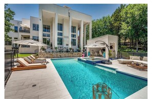 A modern pool area with lounge chairs and umbrellas, a stone wall, and a large building with columns in the background. - Lux Stunning Mansion, Pool. Minutes to AT&T Stadium FIFA, and Six Flags! (Arlington)