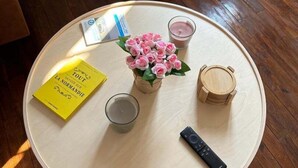 A round wooden table with a yellow book, a glass of water, a vase of pink flowers, a remote control, and a small wooden container. - Idyllic Normandy stay (Veules-les-Roses)