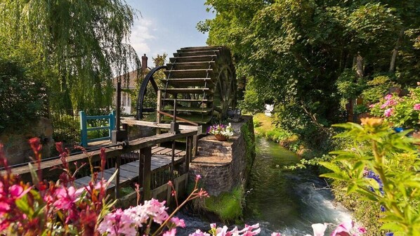 Une structure en bois abritant une roue à eau, avec une plateforme et des marches en bois, le tout entouré d’une végétation luxuriante et traversé par un petit ruisseau.