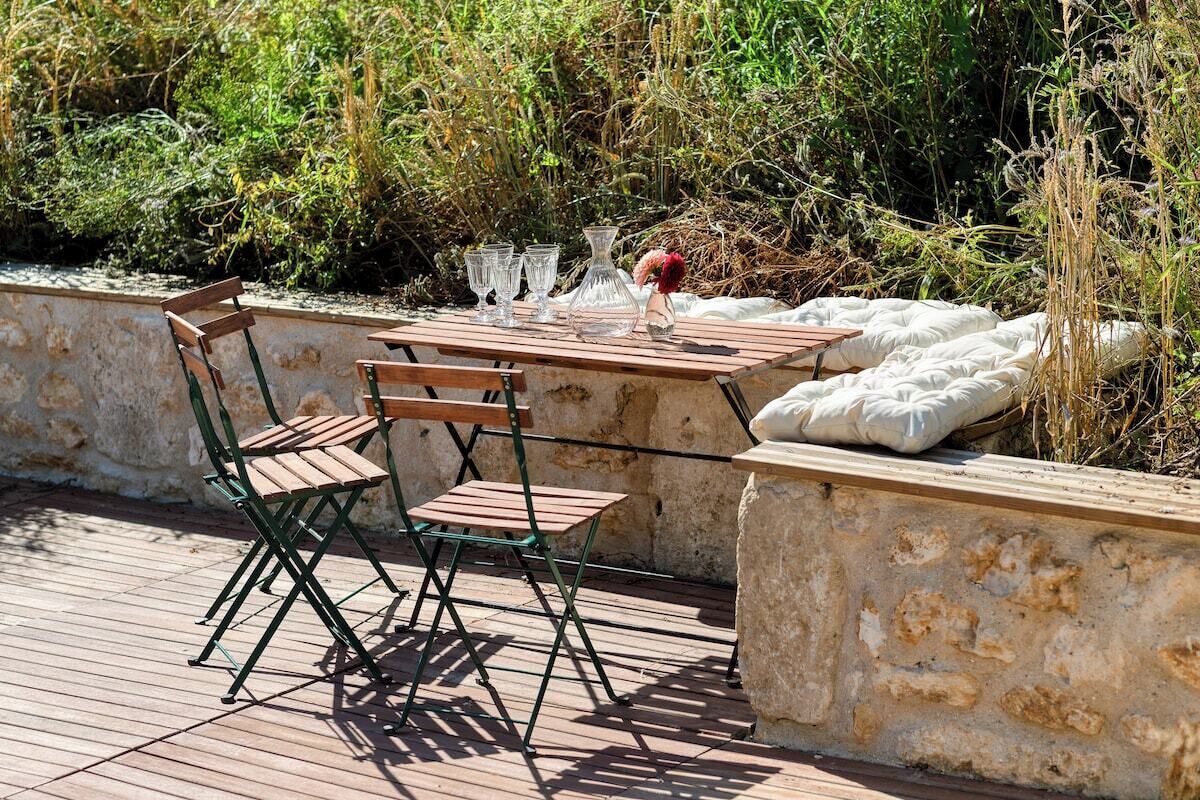 Un área de comedor al aire libre con una mesa y sillas de madera, rodeada de muros de piedra y follaje.