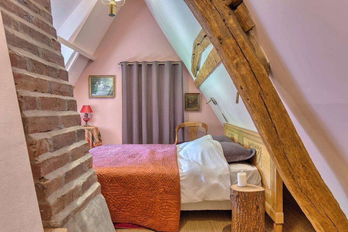 A bedroom with a sloped ceiling, a bed with a white and orange blanket, a wooden headboard, a nightstand with a candle, and a window with curtains.