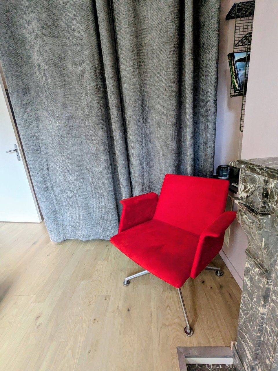Una silla roja en una habitación con suelo de madera, cortinas grises y pared de piedra.