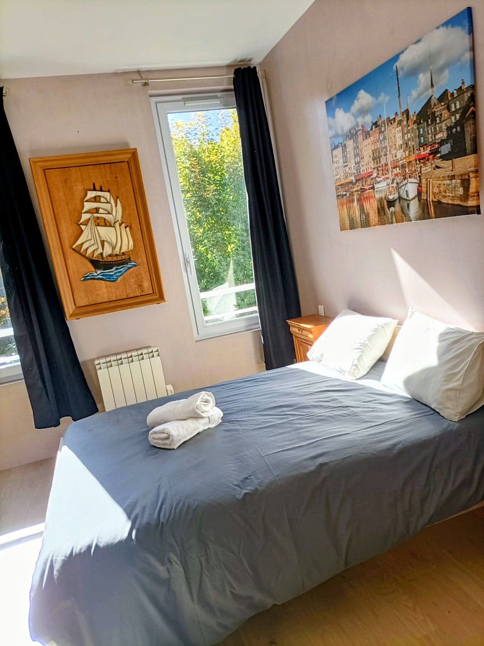 Quarto com cama, janela com cortinas, radiador, armário de madeira com decoração de navio e pintura de paisagem urbana.
