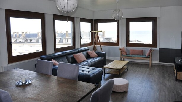Una sala de estar moderna con un sofá de cuero negro, una mesa de centro de madera y una mesa de comedor. Amplias ventanas ofrecen vistas de la ciudad.