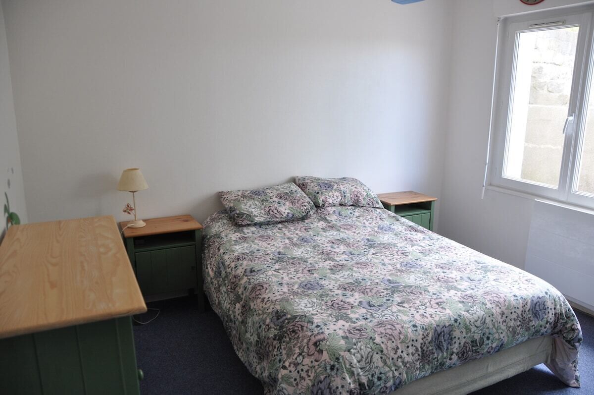 Een slaapkamer met een enkel bed, een houten bureau, een nachtkastje en een raam.