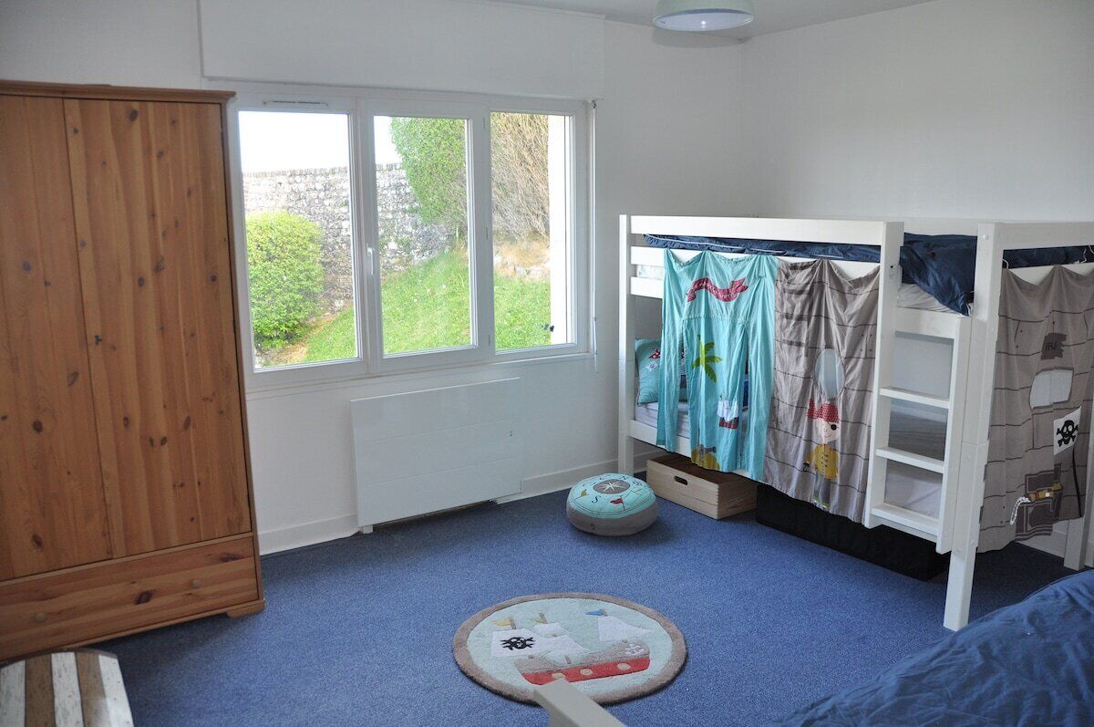 Un dormitorio infantil con literas, un armario de madera, una ventana con vistas a follaje y una alfombra azul.