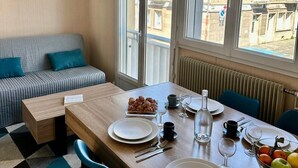 A dining area with a wooden table set for two, a sofa with cushions, and large windows with a view of buildings.