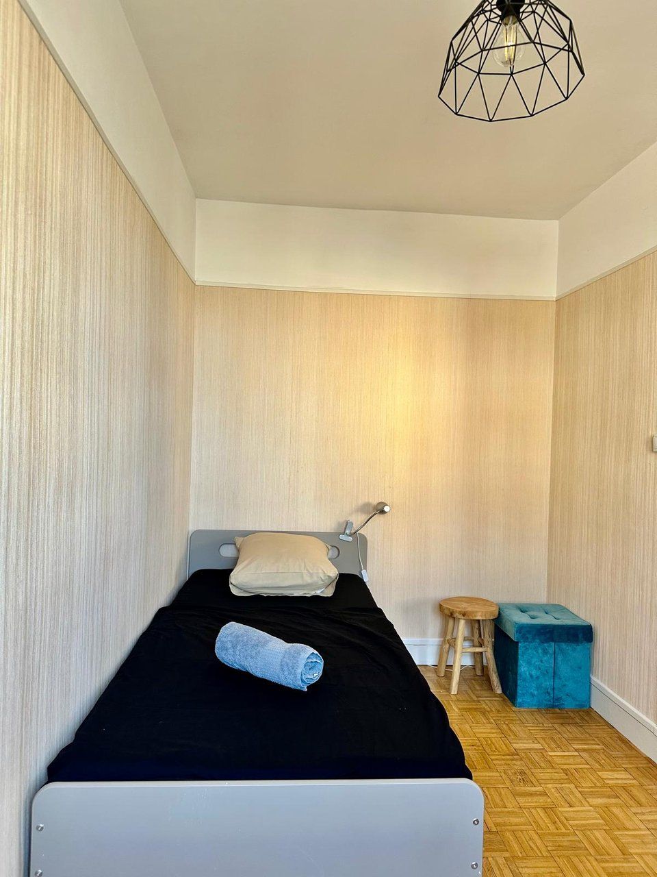 Een kleine, verzorgde slaapkamer met een bed, nachtkastje, krukje en een bijzondere geometrische lamp.