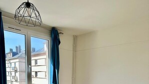 A bedroom with a large bed, a radiator, a window with curtains, and a view of buildings outside.