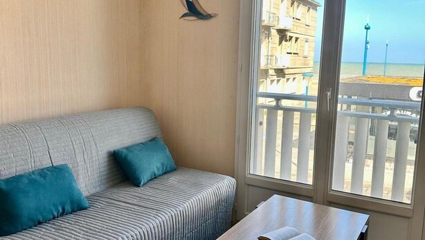 A room with a sofa, a wooden table, and a view of a building and beach through large windows.