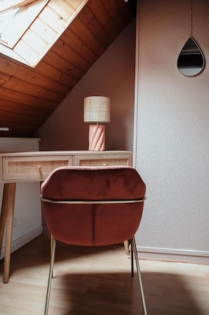 Ein gemütliches Zimmer mit schrägen Wänden, einem Schreibtisch mit Lampe und einem roten Sessel.