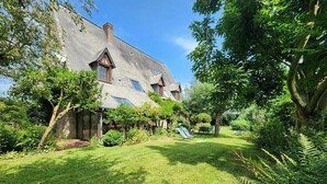 Ein Haus mit geneigtem Dach, einem Ziegelkamin und einem gepflegten Garten mit Rasen, Bäumen und einer Rutsche.
