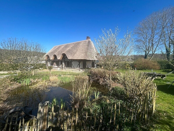 A traditional half-timbered house with a thatched roof, surrounded by a pond and lush greenery.
