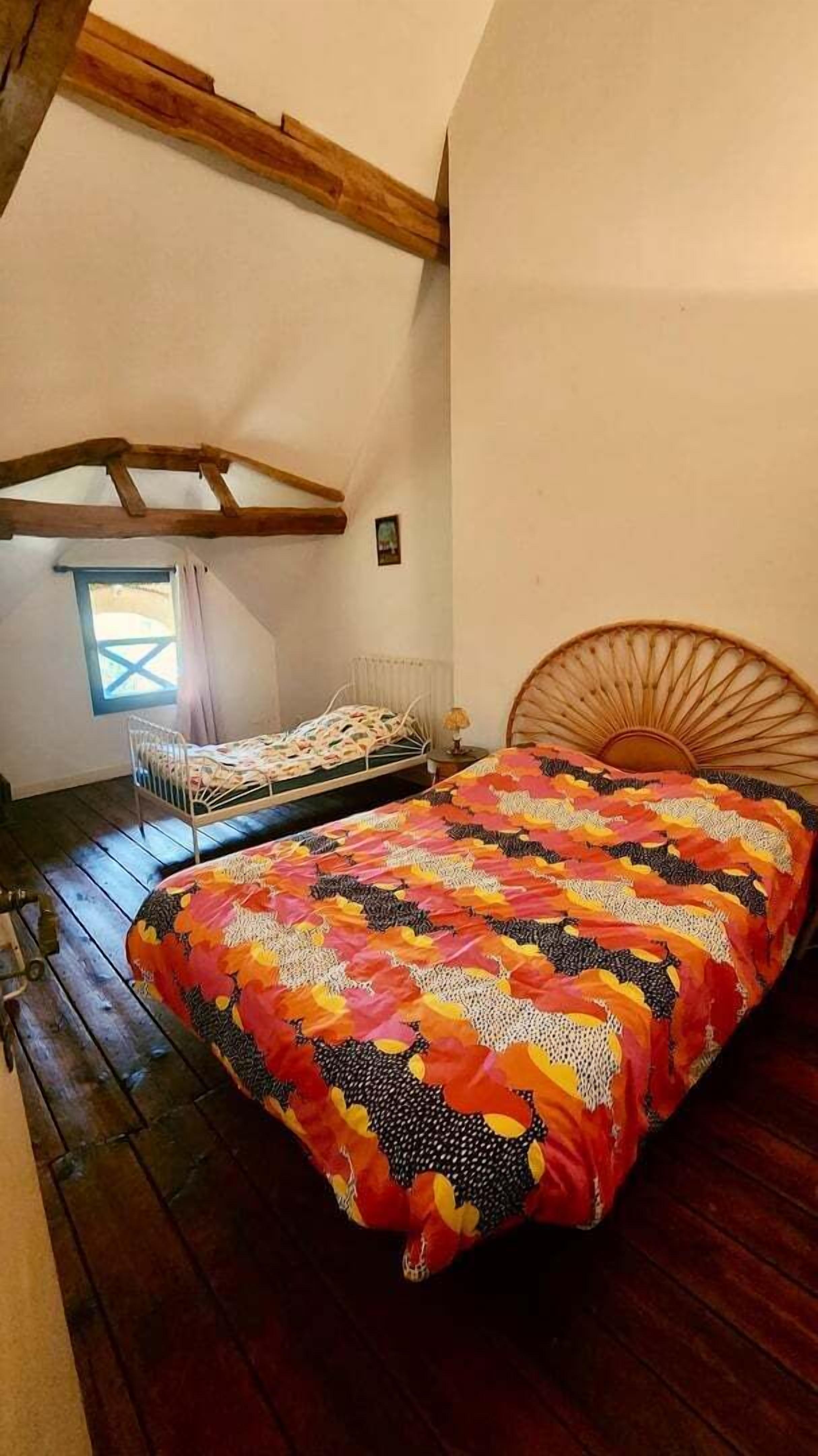 Une chambre avec un plafond en bois, un lit recouvert d’une courtepointe aux couleurs vives, une fenêtre donnant sur un beau paysage et une petite table de chevet.