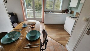 A dining area with a wooden table set for four, a kitchen with white cabinets, and a view through large windows. - Roirand | Romantic Beach on Foot (Veules-les-Roses)