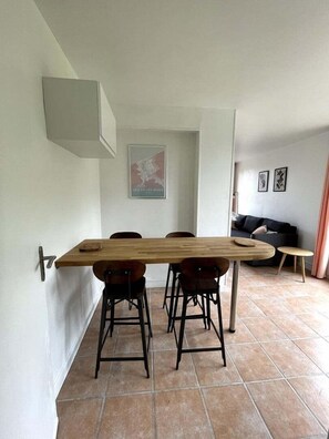 A dining area with a long wooden table, four chairs, and a small round table. There is a sofa and a wall-mounted shelf in the background. - Roirand | Romantic Hideaway in Veules-les-Roses • Beach on Foot (Veules-les-Roses)