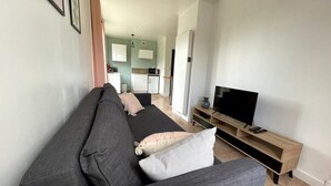 A modern living room with a grey sofa, a TV on a wooden stand, and a kitchen in the background. - Roirand | Romantic Beach on Foot (Veules-les-Roses)