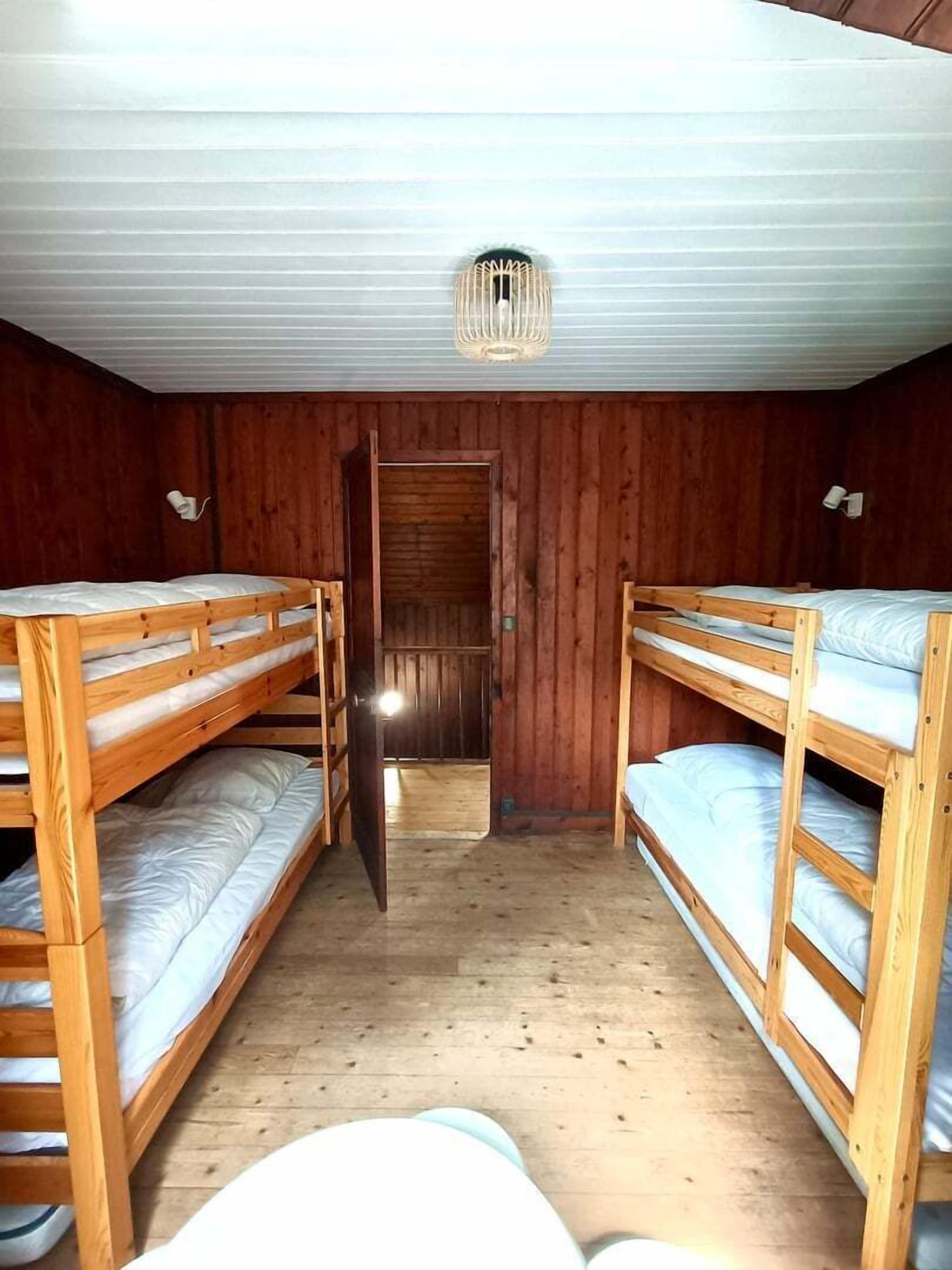 A room with bunk beds, wooden walls, a ceiling light, and a door leading to another room.