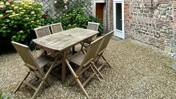 Une cour avec une table et des chaises en bois, entourée d’un sol en gravier et d’un mur en briques.