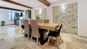 Ein Essbereich mit Holztisch und gewobenen Stühlen, eine Küche im Hintergrund und eine Wand aus strukturiertem Stein.