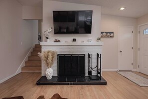 Una sala de estar con chimenea, televisor montado encima y una escalera que sube a otro nivel.