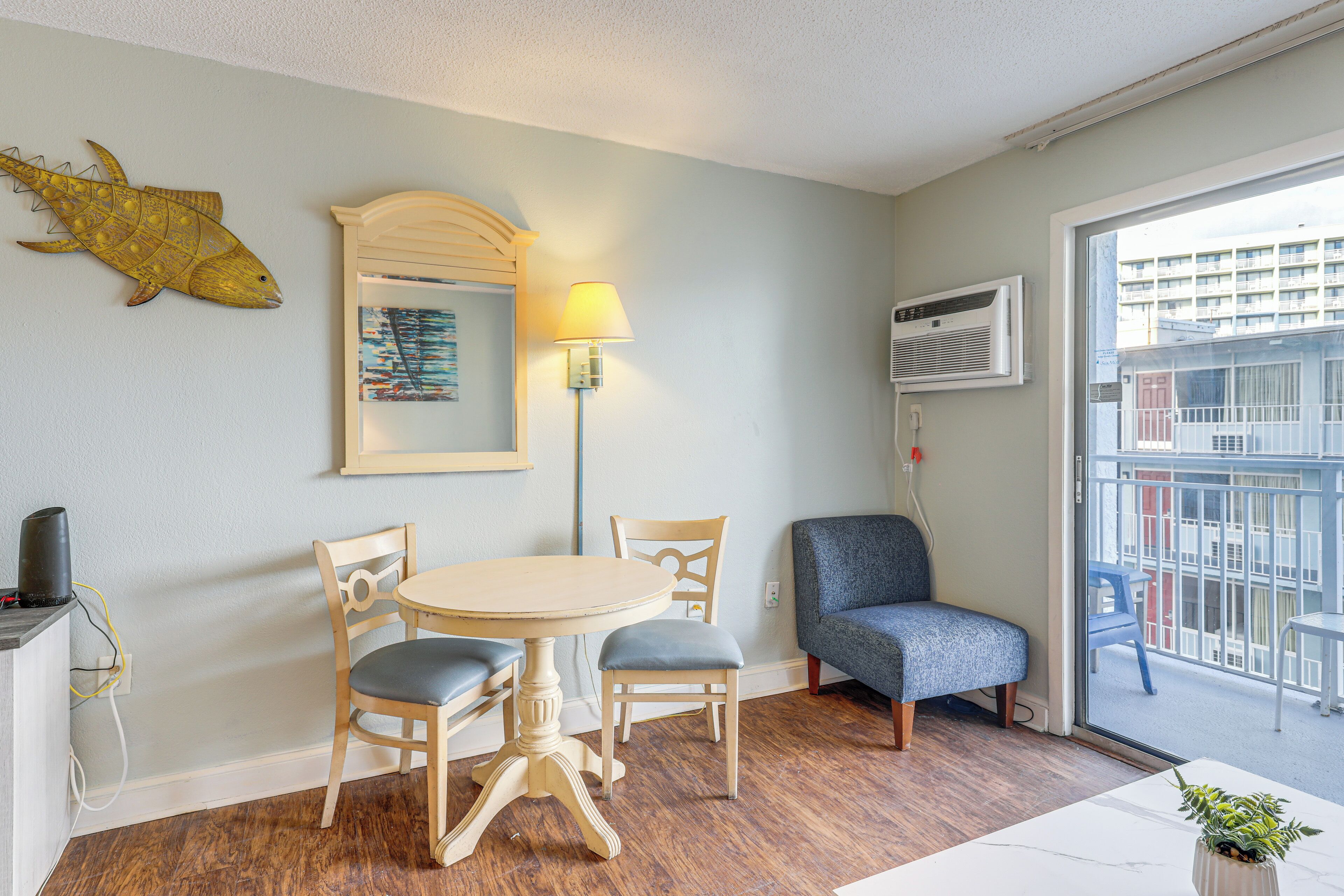 A room with a round table, two chairs, a lamp, a fish decoration, and a view of buildings through a balcony door.