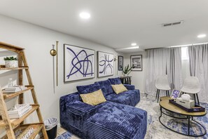 A modern living room with a blue patterned sofa, a wooden bookshelf, and abstract wall art. - Walk to Patterson Park! Pet-Friendly Townhome (Baltimore)