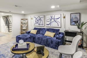 A modern living room with a blue sofa, a grey armchair, and a coffee table. There are abstract paintings on the wall and a staircase leading to another room. - Walk to Patterson Park! Pet-Friendly Townhome (Baltimore)