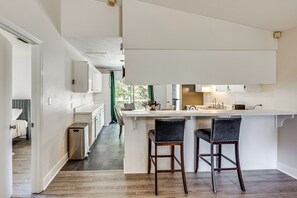 Eine moderne Küche mit einer zentralen Kochinsel, zwei Barhocker und Blick in das angrenzende Schlafzimmer.