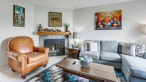 A living room with a leather armchair, a grey sofa, a fireplace, and various paintings on the walls.
