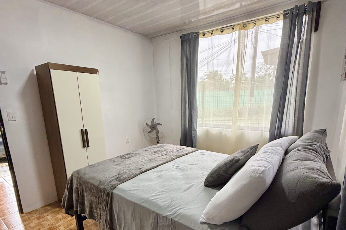 A bedroom with a bed, a wardrobe, a fan, a window with curtains, and a mat labeled 'HOME' on the floor.