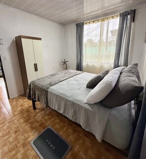 A bedroom with a bed, a wardrobe, a fan, a window with curtains, and a mat labeled 'HOME' on the floor. - Apartamento Lapas  (La Fortuna)