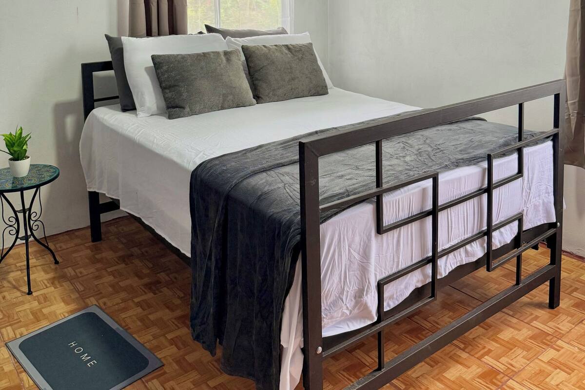 A bedroom with a bed, a small table with a plant, a window with curtains, and a radiator.