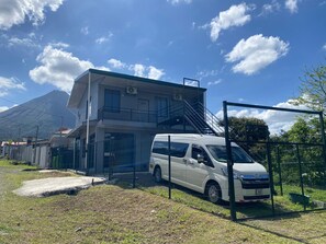 A two-story building with a staircase leading to the upper floor, a fenced parking area, and a mountain in the background. - Apartamento Lapas  (La Fortuna)