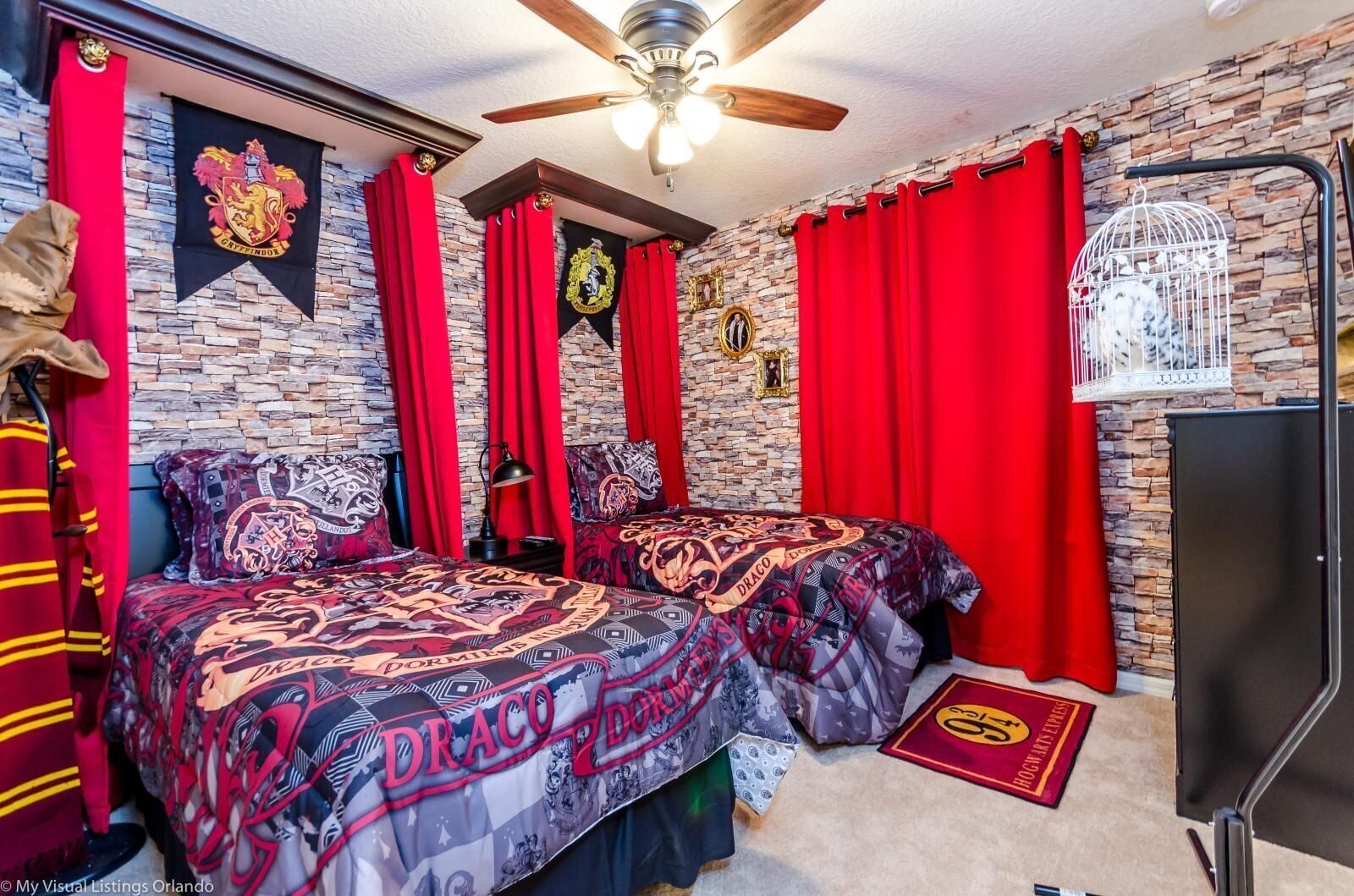Une chambre avec deux lits, des rideaux rouges, un ventilateur de plafond, un mur en pierre et les armoiries de Poudlard.