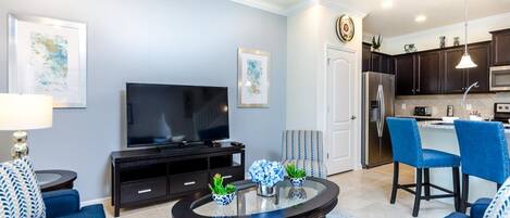 A modern living room with a flat-screen TV, a glass coffee table, and blue sofas.