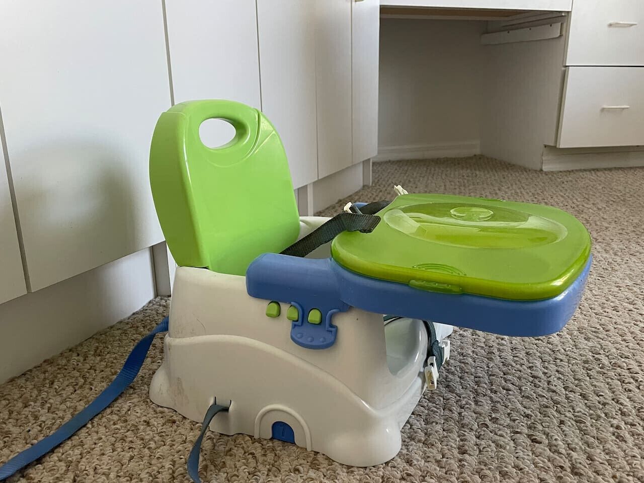 Kannettava WC, jossa on vihreä istuin ja sininen kahva, on sijoitettu matolla päällystetylle lattialle huoneeseen, jossa on valkoiset kaapit.