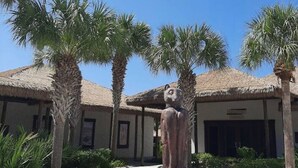 Eine Skulptur einer sitzenden Figur auf einem Sockel in einem Innenhof mit Palmen und einem Gebäude mit einem Strohdach.