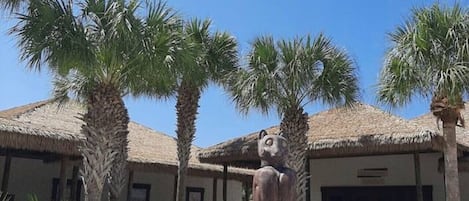 Een sculptuur van een zittend figuur op een sokkel in een binnentuin met palmboompjes en een gebouw met rieten dak.