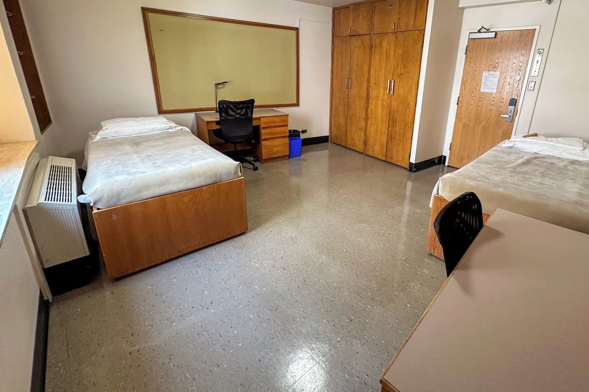 Quarto de estudante com duas camas, uma escrivaninha, uma cadeira, um guarda-roupa e um radiador.
