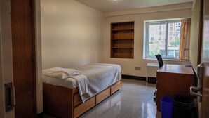 Un dormitorio con una cama, un escritorio, una silla, una ventana y un cubo de basura azul.