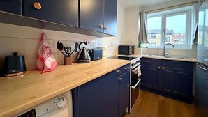 A modern kitchen with dark blue cabinets, a wooden countertop, a washing machine, a microwave, and a window with a view of buildings outside. - Cosy 1BR Flat in East London (London)
