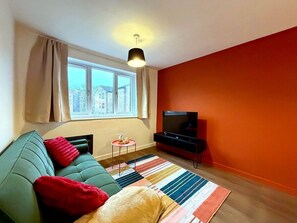 A modern living room with a green sofa, a red and beige blanket, a striped rug, and a TV on a black stand. - Cosy 1BR Flat in East London (London)