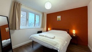 A bedroom with a bed, a window with curtains, a bedside lamp, and a mirror. - Cosy 1BR Flat in East London (London)
