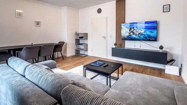 A modern living room with a sofa, a coffee table, a TV, and a dining area with chairs. - Modern & Cozy Apartment | Central Magdeburg | (Magdeburg)