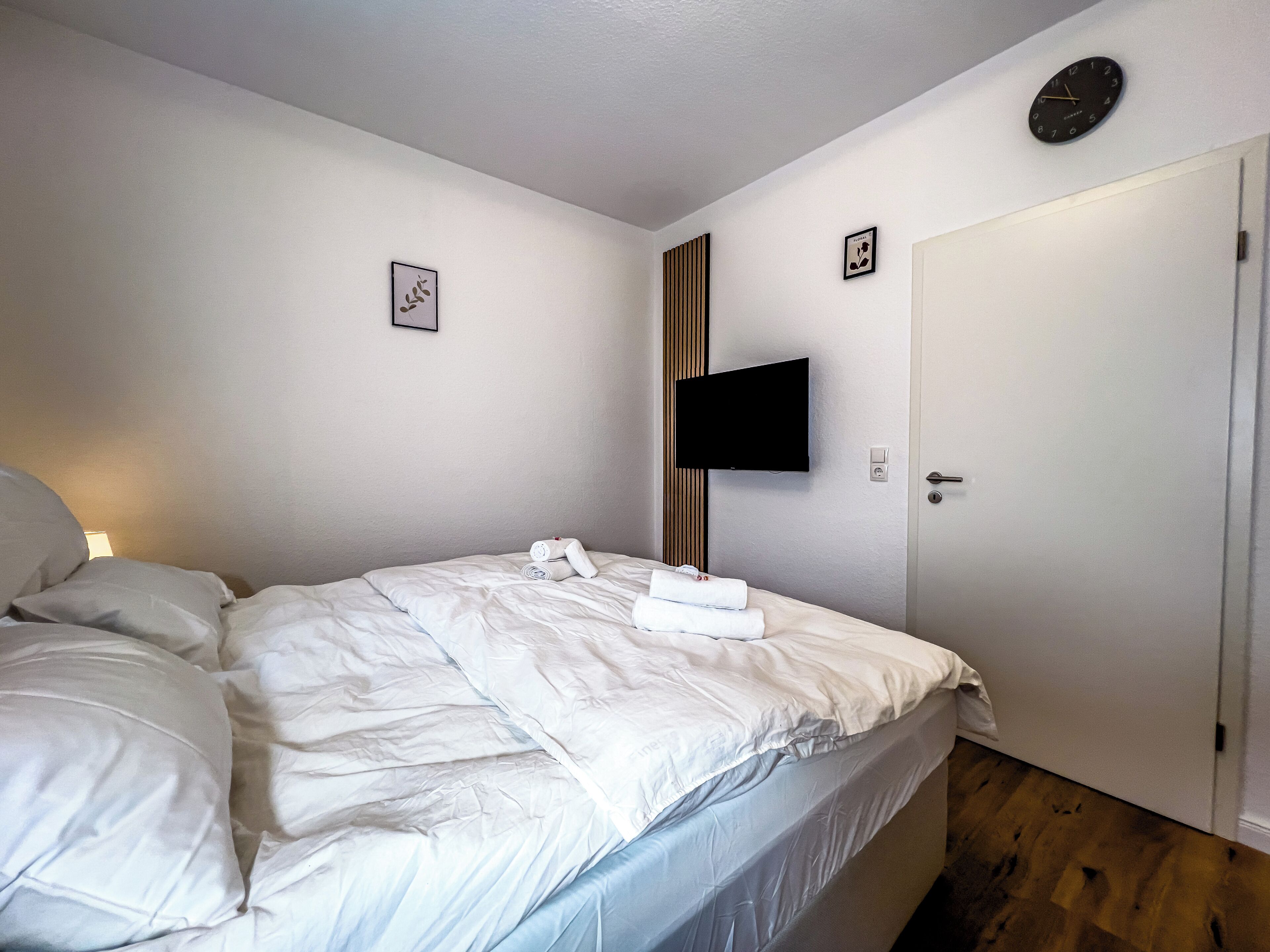 Een netjes opgemaakt bed met witte lakens, een flatscreen televisie aan de muur bevestigd en een deur met een handvat.