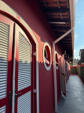 A row of red and white doors with shutters, a clear sky, and a brick wall in the background. - Chalés Rosamar (Guarujá)