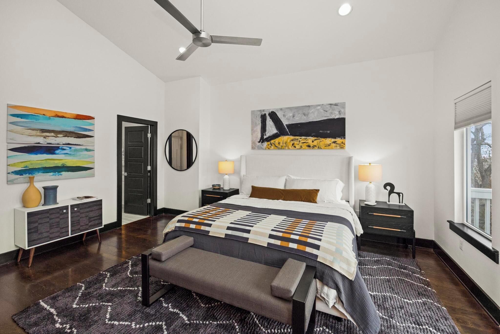 Une chambre à coucher moderne avec un grand lit, des tables de chevet, un ventilateur de plafond et des œuvres d’art accrochées aux murs.